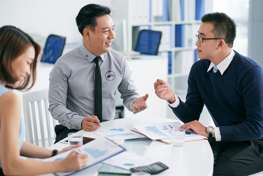 Three people having a conversation over a table with documents, two glasses of water, and a calculator on top of it.