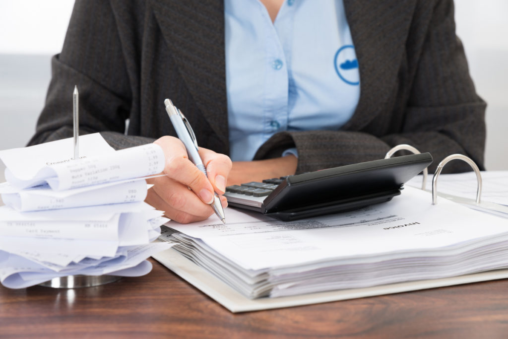 A female professional writing on a stack of documents with a calculator and receipts pinned on the desk she is using.