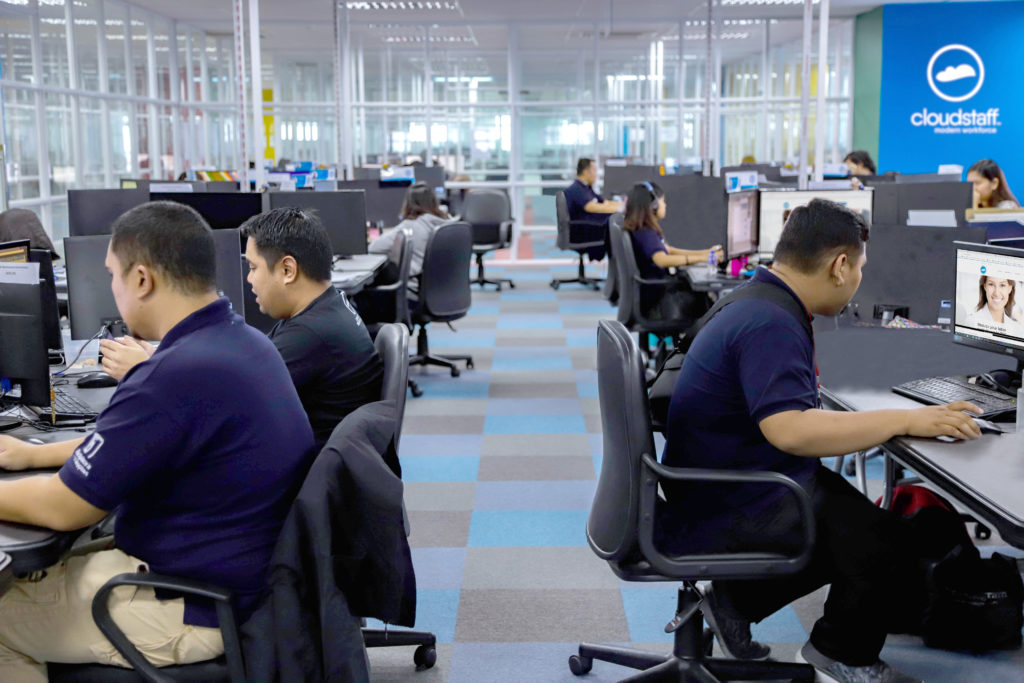Employees working in a production area. All of them face their computers and their background shows the Cloudstaff logo.
