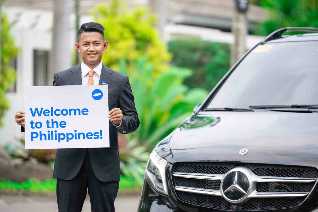 A man in suit holding a signage that reads "Welcome to the Philippines" beside a car.