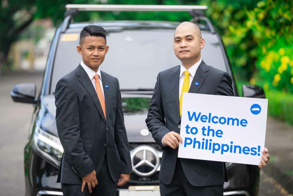 Two men in suit holding a signage that reads "Welcome to the Philippines" in front of a car.